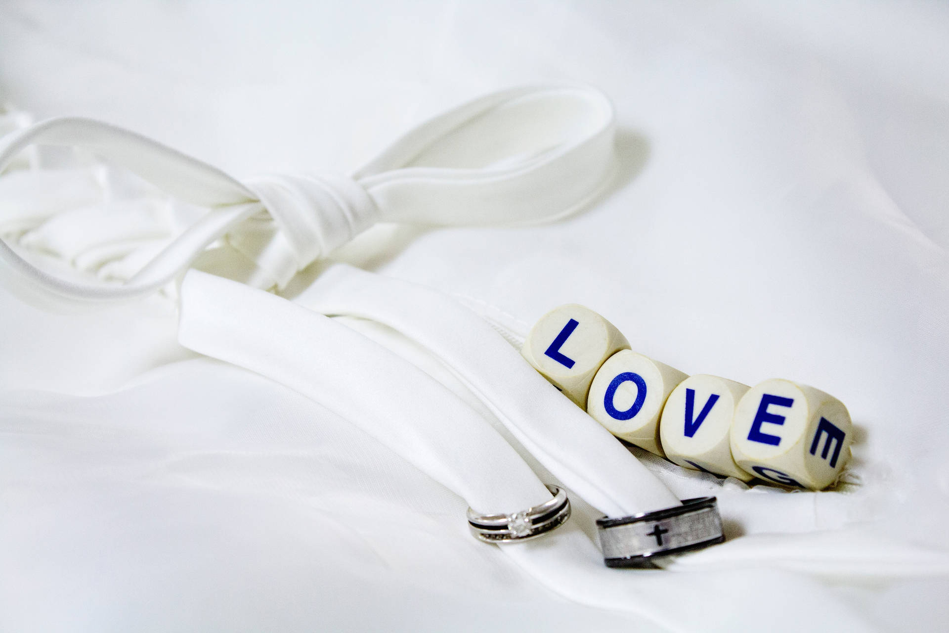 চারটি সাদা পাশার উপর 'Love' লেখা, যার পাশে একটি সাদা ফিতা দিয়ে বাঁধা দুটি রূপালী ও কালো বিবাহের আংটি একটি সাদা চাদরের উপর রাখা। — ভালোবাসা ওয়ালপেপার